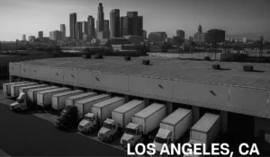 Black and white photograph of a distribution center in Los Angeles, CA with delivery trucks backed into loading bays and the downtown skyline in the background.